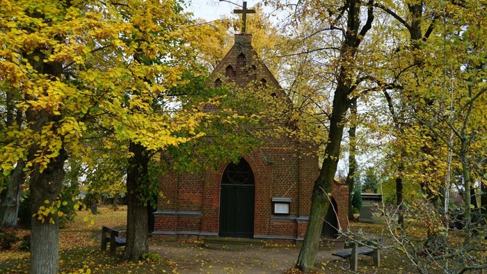 Kleine Backsteinkapelle mit Kreuz auf dem Dach, umgeben von Bäumen mit herbstlich gelben Blättern.