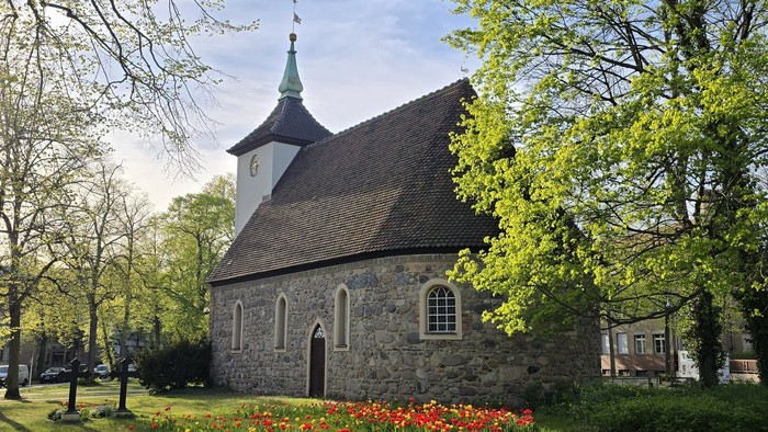 Kleine Kirche mit spitzem Turm, umgeben von Bäumen und blühenden Blumen