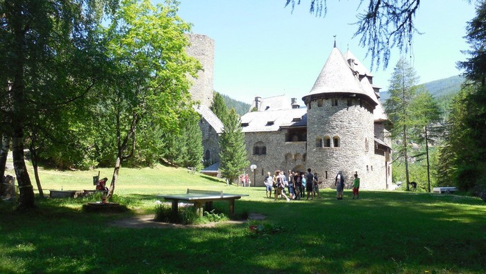 Ein Steinhaus mit Turm, umgeben von Bäumen und Menschen auf der Wiese