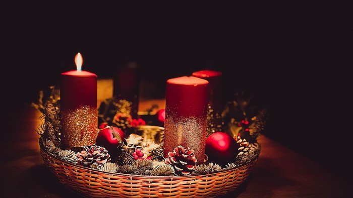 Tre tændte stearinlys i en dekorativ kurv med fyrrekogler og bær.