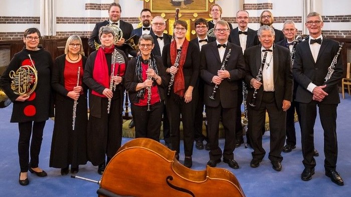 Et formelt orkesterensemble poser sammen med forskellige træblæsere, messingblæsere og strygeinstrumenter i en stor sal.