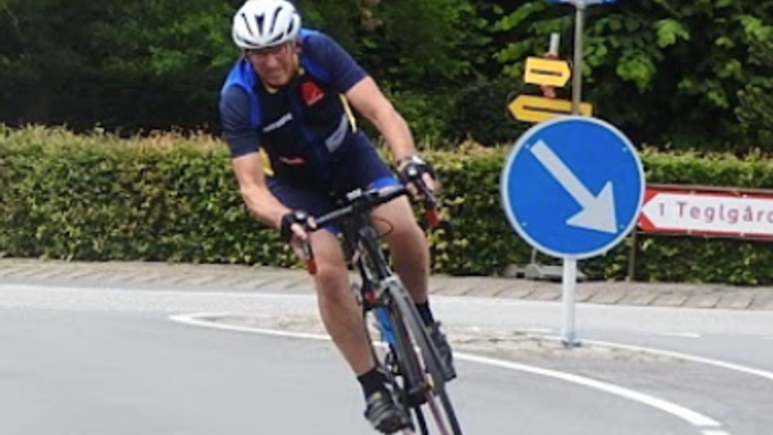 En cyklist i blåt cykeltøj cykler i et sving. I baggrunden kan man se et vejskilt, hvor der står Teglgård 1.