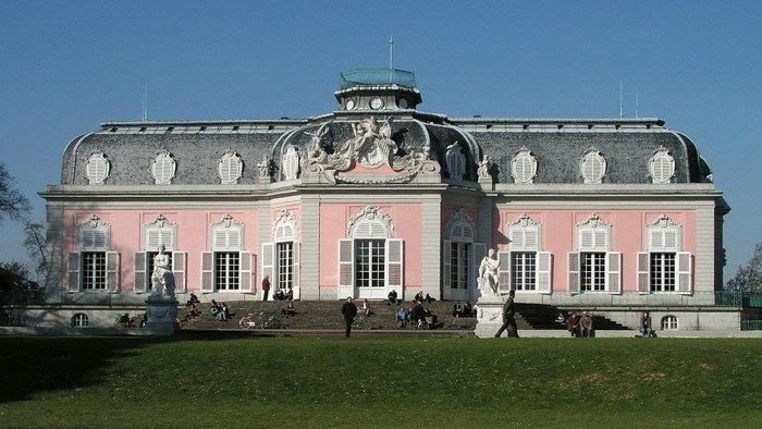 Großes, prächtiges Gebäude mit rosa Fassade und zahlreichen Statuen, umgeben von grünem Rasen und einigen Menschen.