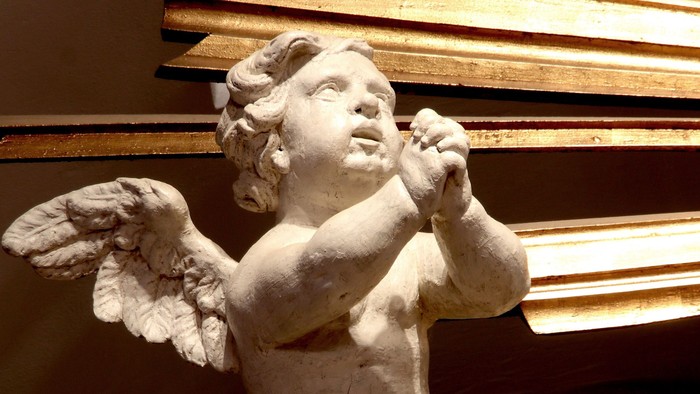 En lille engelstatue med vinger og hånd ved munnen, placeret foran en guldfarvet væg.