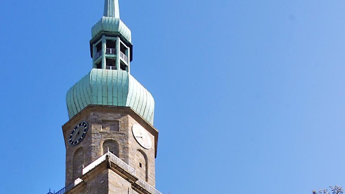 Hohe Kirche mit grünem Turm und Kreuz, umgeben von Bäumen und Menschen