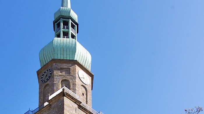 Hohe Kirche mit grünem Turm und Kreuz, umgeben von Bäumen und Menschen