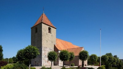 En lille kirke med tårn og røde tag, omgivet af træer og buske under et klarblåt sky.