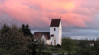 En hvid kirke med tårn og spiret mod en rosa skyer og solnedgang