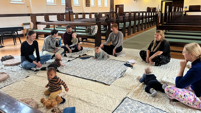 En gruppe mennesker og børn samles indendørs på måtter i en rummelig sal med træbænke.