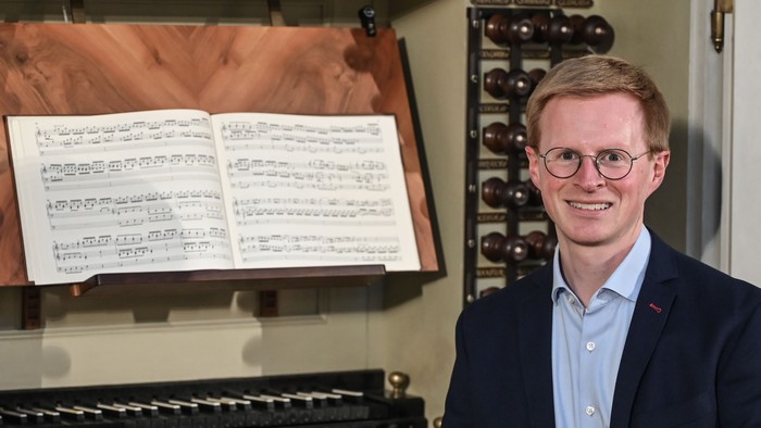 Ein Mann im Anzug lächelt neben einem Flügel, auf dessen Notenständer Notenblätter liegen.