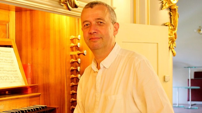 Ein Mann in einem weißen Hemd posiert selbstbewusst neben einer verzierten Pfeifenorgel.