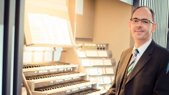 Ein Mann im Anzug sitzt an einer großen Pfeifenorgel und lächelt in die Kamera.