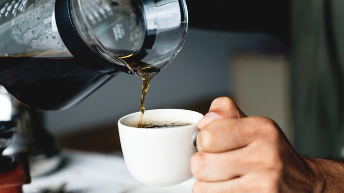 En hånd porer kaffe fra en silverfarvet kaffekanner i en hvid tønde.