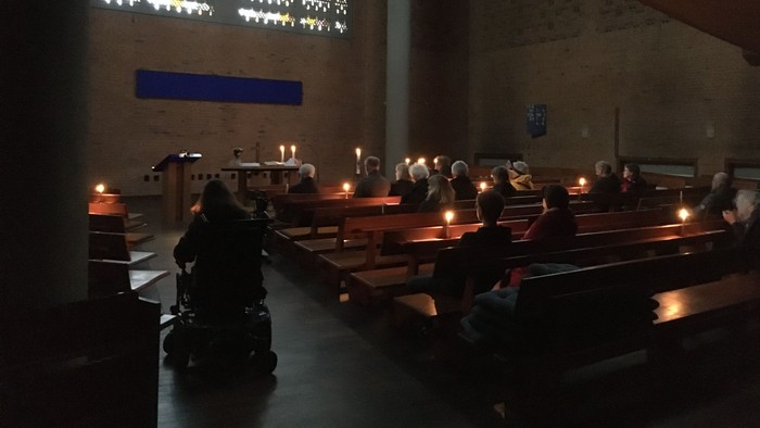 Ein schwach beleuchteter Kircheninnenraum mit Gläubigen, die in den Bänken sitzen, brennenden Kerzen und bunten Glasfenstern.