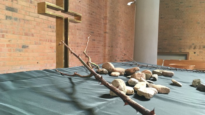 Ein feierlicher Altar, bedeckt mit grünem Tuch, dornigen Zweigen, Steinen und einem Kreuz vor Backsteinwänden.