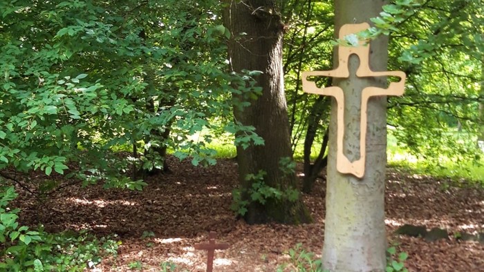 Ein ruhiges Gartendenkmal mit einem hölzernen Kreuz, das an einem Baumstamm befestigt ist, und einem kleinen steinernen Altar darunter.
