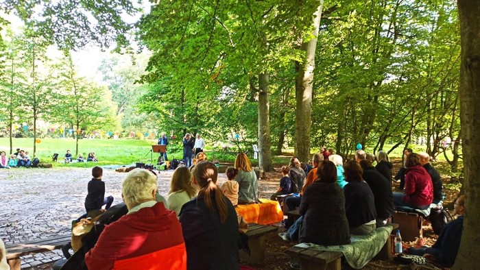 Eine lebhafte Zusammenkunft im Freien, bei der Menschen auf Bänken in einem üppigen, grünen Park aufmerksam zuhören.