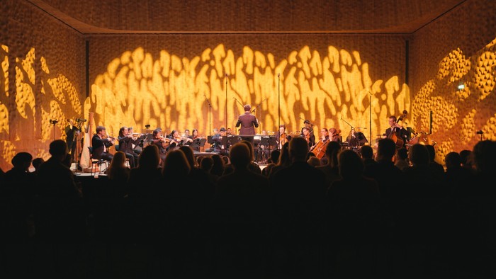 Ein Orchester spielt auf einer Bühne vor einem beleuchteten Hintergrund.