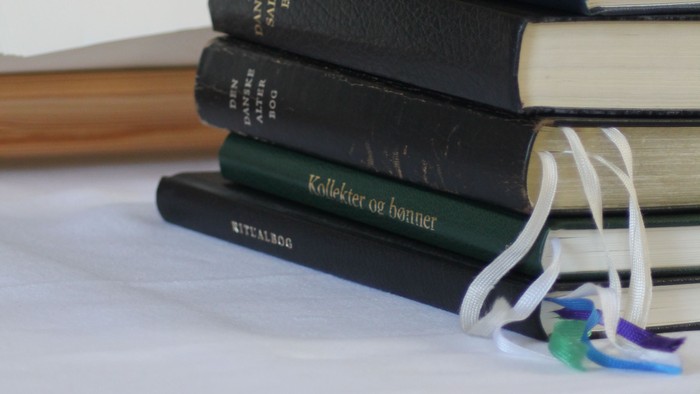 En stabel af bøger på seng, med en åben bog i baggrunden