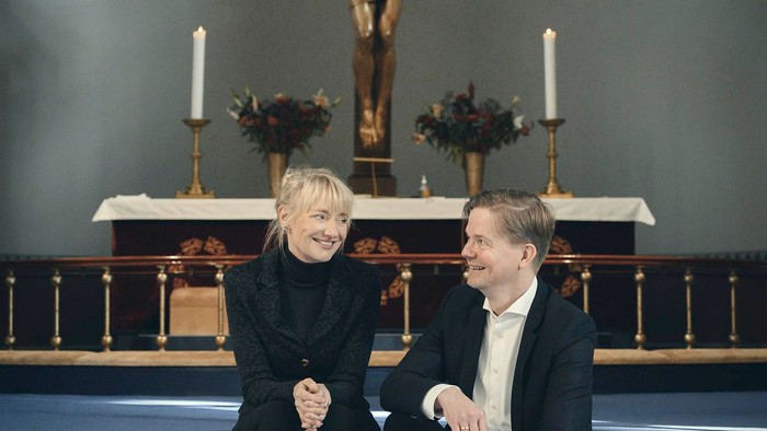 En mand og en kvinde sidder i en kirke foran alteret med en korsfæstet Jesus.