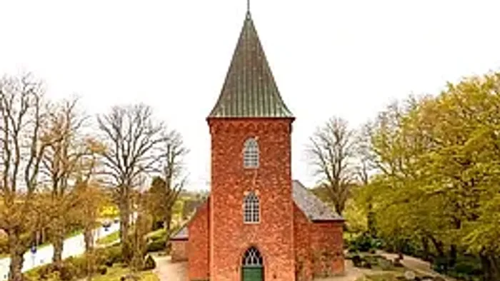 Kleine rote Kirche mit hohem Turm in parkähnlichem Garten.