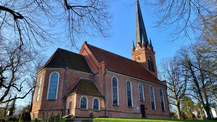 Kirchengebäude aus Ziegeln mit hohem Turm und spitzen Fenstern, umgeben von Bäumen und grünem Gras