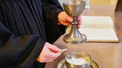 Pfarrer im Talar hält einen Kelch und eine Hostie über einer Schale mit weiteren Hostien auf einem Altar.