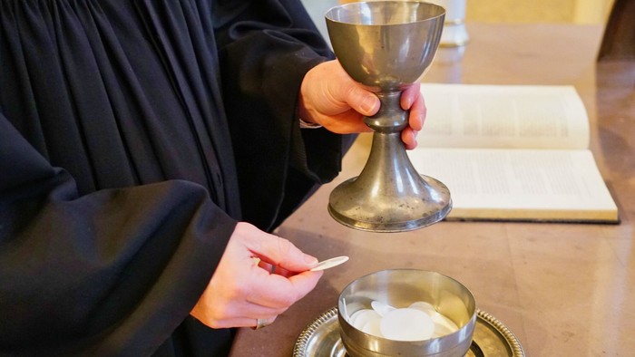 Pfarrer im Talar hält einen Kelch und eine Hostie über einer Schale mit weiteren Hostien auf einem Altar.