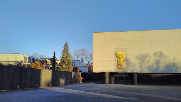 Großes weißes Gebäude mit goldenem Emblem, umgeben von Bäumen und Gehweg