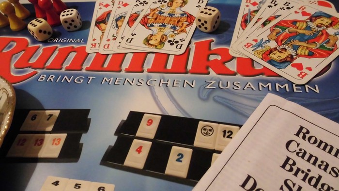Eine Tischansicht des Kartenspiels Rommé mit Würfeln, Karten, Zählsteinen und Spielregeln.