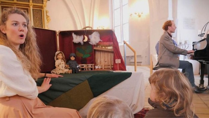 En kvinde læser højt for børn, mens en mand spiller klaver i et elegant rum.