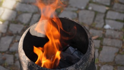 Ein rustikaler Feuerkorb brennt hell auf einem Kopfsteinpflaster, während Flammen darin flackern.