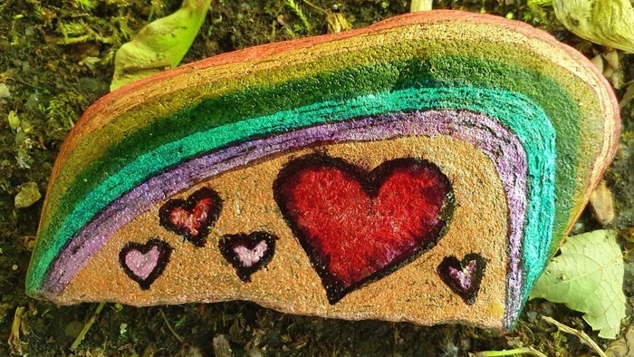 Bemalter glatter Stein mit Regenbogenstreifen, rotem Herz und kleineren Herzen in schwarzer Umrisslinie auf natürlichem Untergrund.
