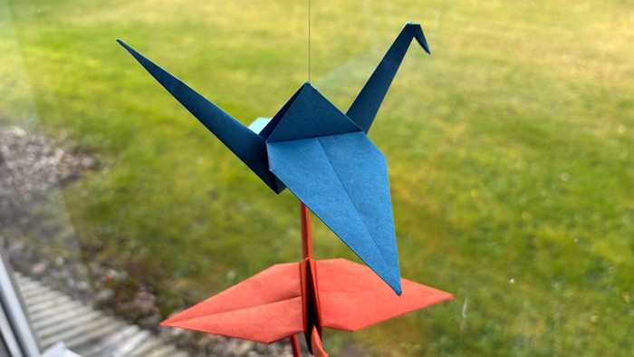 Drei Origami-Vögel hängen an einem Faden vor einem Fenster mit Blick auf den Garten.