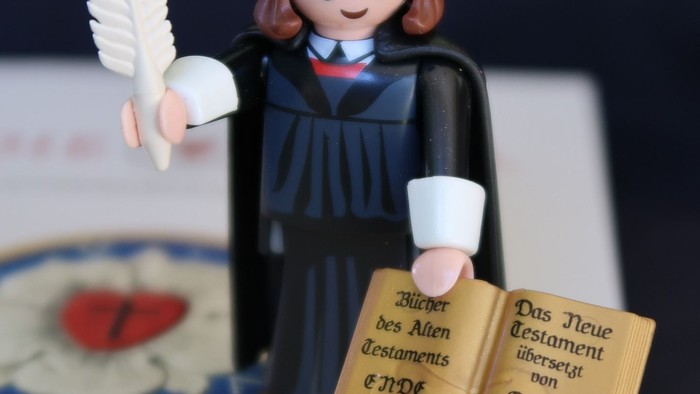 Eine Playmobil-Figur in Doktorrobe hält eine Feder und ein aufgeschlagenes Buch mit Text von Martin Luther.