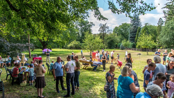 Eine Gruppe von Menschen feiert im Freien, einige sitzen an Tischen, andere stehen und unterhalten sich, umgeben von Bäumen und Gras.