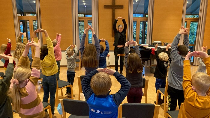 Kinder in einem Kirchenraum heben die Hände während einer Gruppenaktivität, die von einer erwachsenen Person geleitet wird.