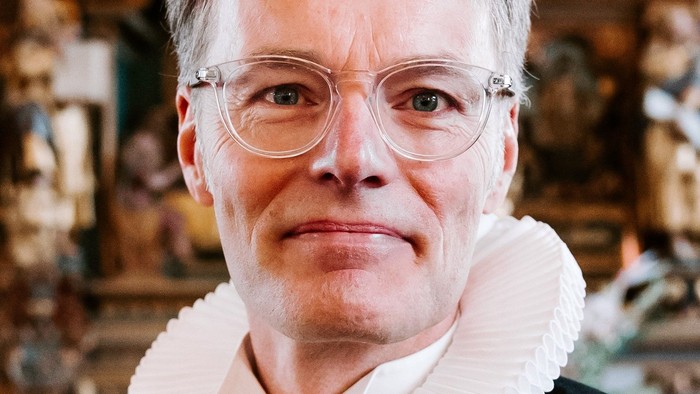 En kvinde med hvid krage og briller står foran en prægtig og udsmykket baggrund