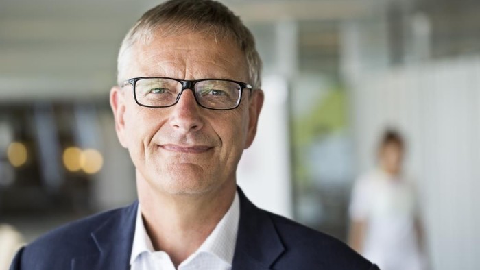 En mand i blå jakke og briller smiler i et kontor.