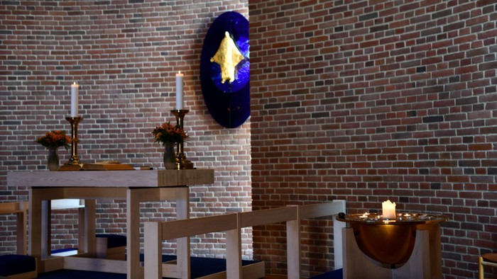 En lille kapel med mur af røde mursten, med bord og stol, lysstake og blå skjold med stjerne.