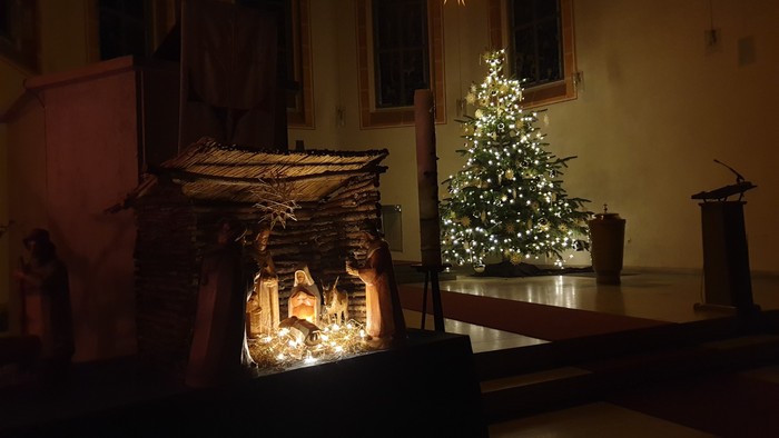 In der Kirche steht ein beleuchteter Weihnachtsbaum neben einer Krippe.
