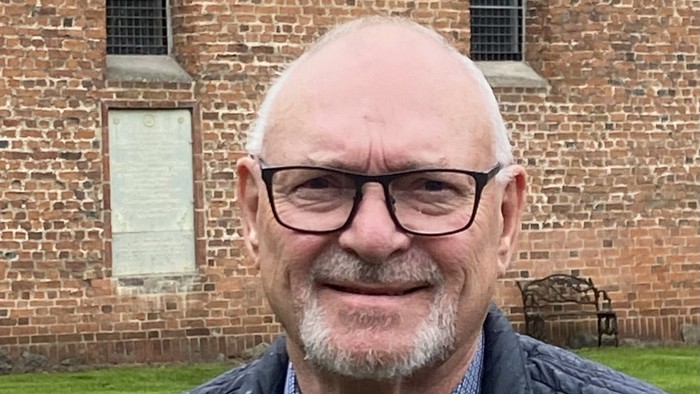 En smilende, bald, ældre mand med briller og blå vest over et skjorte står foran en mur af røde mursten.