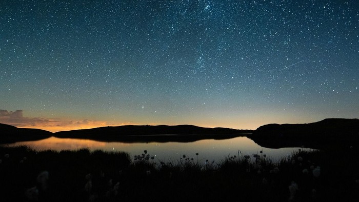 Sternenklarer Nachthimmel spiegelt sich über einem ruhigen See, umgeben von silhouettierten Hügeln.