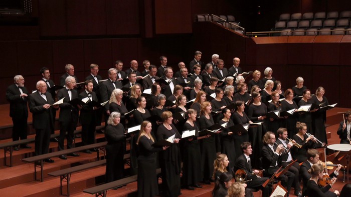 Ein großer Chor und ein Orchester, die gemeinsam in einem Konzertsaal auftreten.