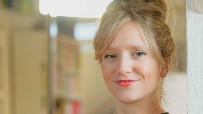Eine Frau mit blondem Haar steht vor einem Bücherregal.