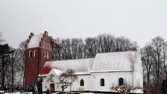 En snedækket kirke med et rødt tårn og et hvidt tag, omgivet af bærbare træer og sne.