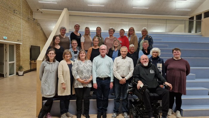 En gruppe mennesker, der poser til et foto – nogle står, nogle sidder i kørestole – i et rummeligt lokale med trappe.