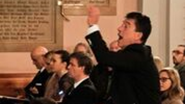 Ein Chor probt in einem großen Raum, geleitet von einem Dirigenten.