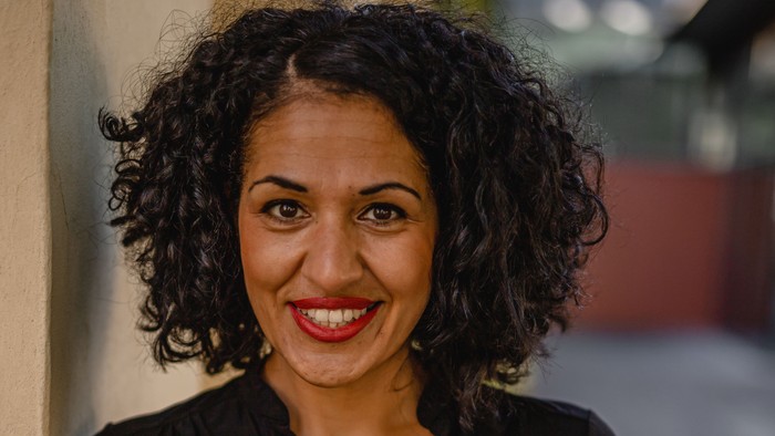 Lächelnde Frau mit lockigem Haar in einem schwarzen Oberteil, die draußen steht.