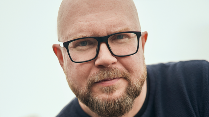 Skaldet mand med briller og skæg iført en mørkeblå skjorte.